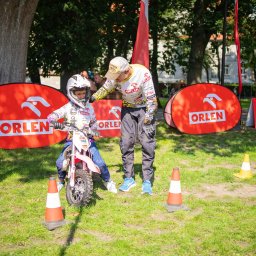 ArtGate - Młody motocyklista w kasku na małym motocyklu, instruowany przez dorosłego mężczyznę na trawiastym terenie z pachołkami i banerami Orlen w tle.