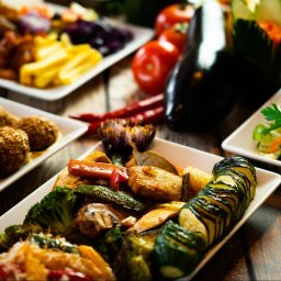 ArtGate - Profesjonalna fotografia kulinarnej kompozycji na drewnianym stole w Warszawie, prezentująca różnorodne dania wegetariańskie w białych naczyniach, w tym falafel z sezamem, grillowane warzywa...