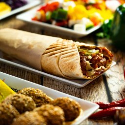 ArtGate - Shawarma w papierze na białym talerzyku, obok pomidory, papryczki chili, falafel i sałatki na drewnianym stole.