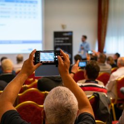 ArtGate - Osoba fotografuje slajd prezentacji z diagramem na ekranie smartfona podczas konferencji, widoczni inni uczestnicy również robiący zdjęcia.