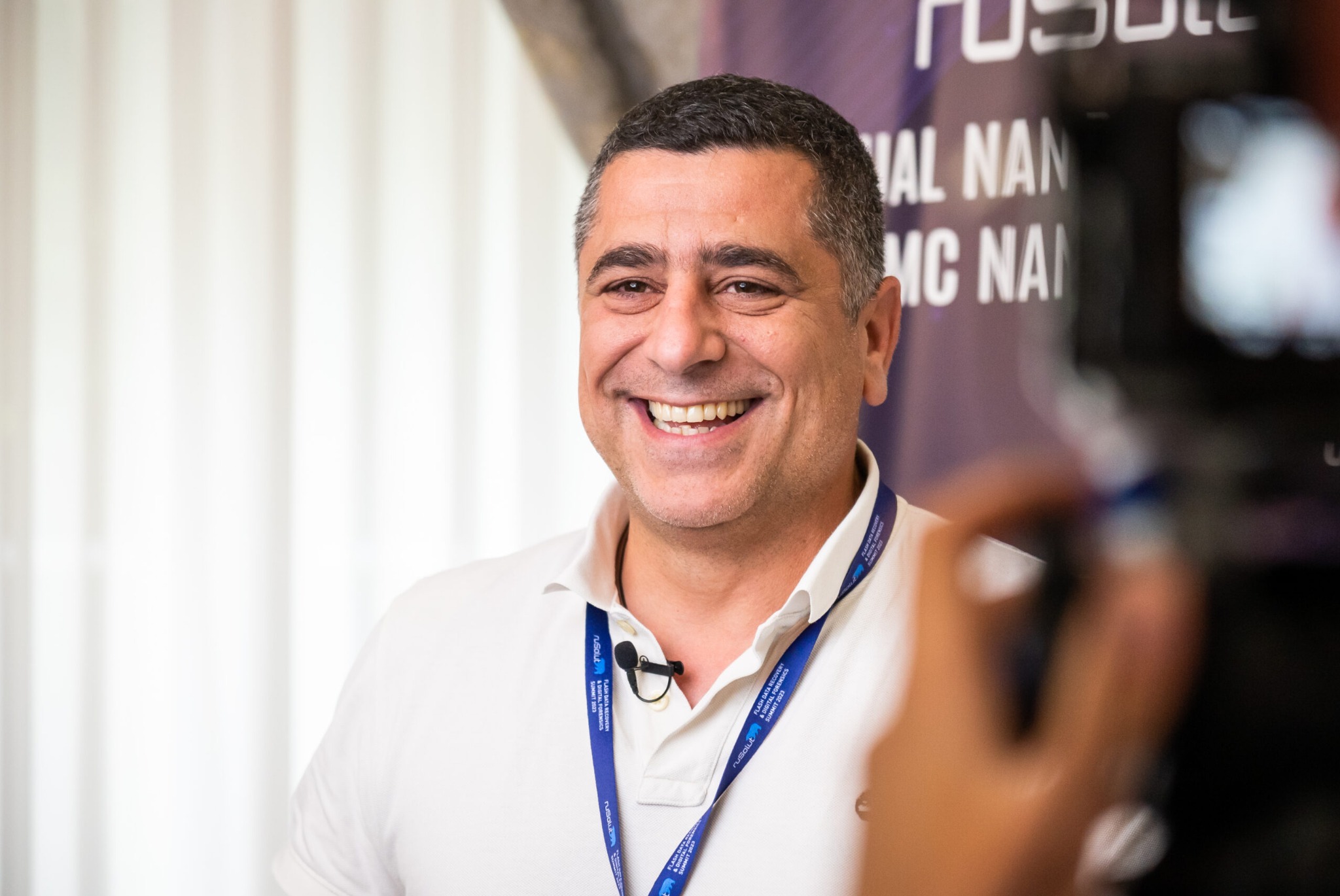 Uśmiechnięty mężczyzna z mikrofonem przypiętym do koszuli polo, nagrywany kamerą podczas wydarzenia branżowego.