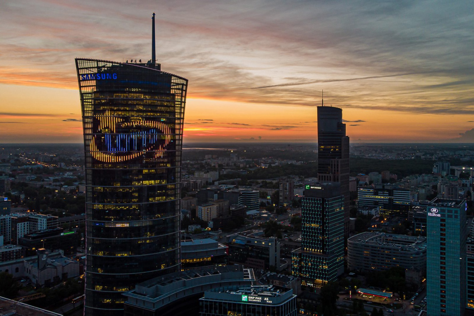 Warszawa nocą: wysoki budynek z logo Samsung i wyświetlanym znakiem graficznym na elewacji, w tle zachodzące słońce i panorama miasta.