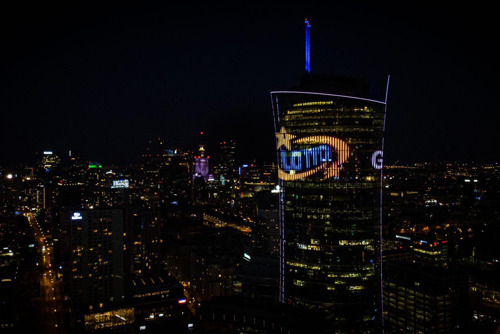 Nocny widok na Warszawę z podświetlonym wieżowcem Warsaw UNIT z logo LOTTO, w tle Pałac Kultury i Nauki oraz inne budynki miasta.