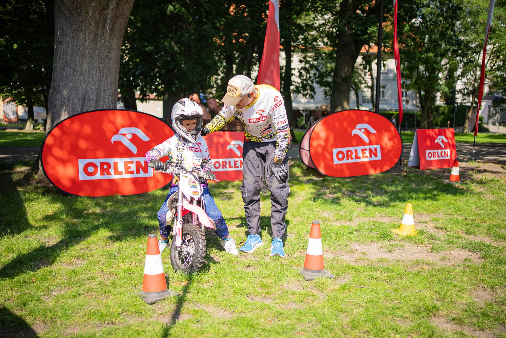 Młody motocyklista w kasku na małym motocyklu, instruowany przez dorosłego mężczyznę na trawiastym terenie z pachołkami i banerami Orlen w tle.