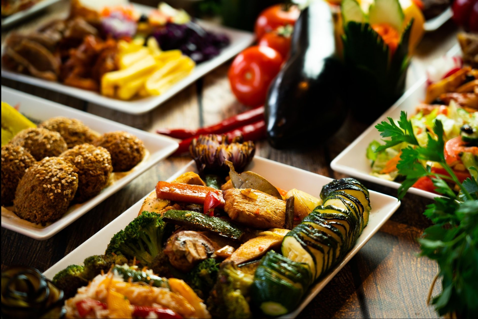 Profesjonalna fotografia kulinarnej kompozycji na drewnianym stole w Warszawie, prezentująca różnorodne dania wegetariańskie w białych naczyniach, w tym falafel z sezamem, grillowane warzywa...