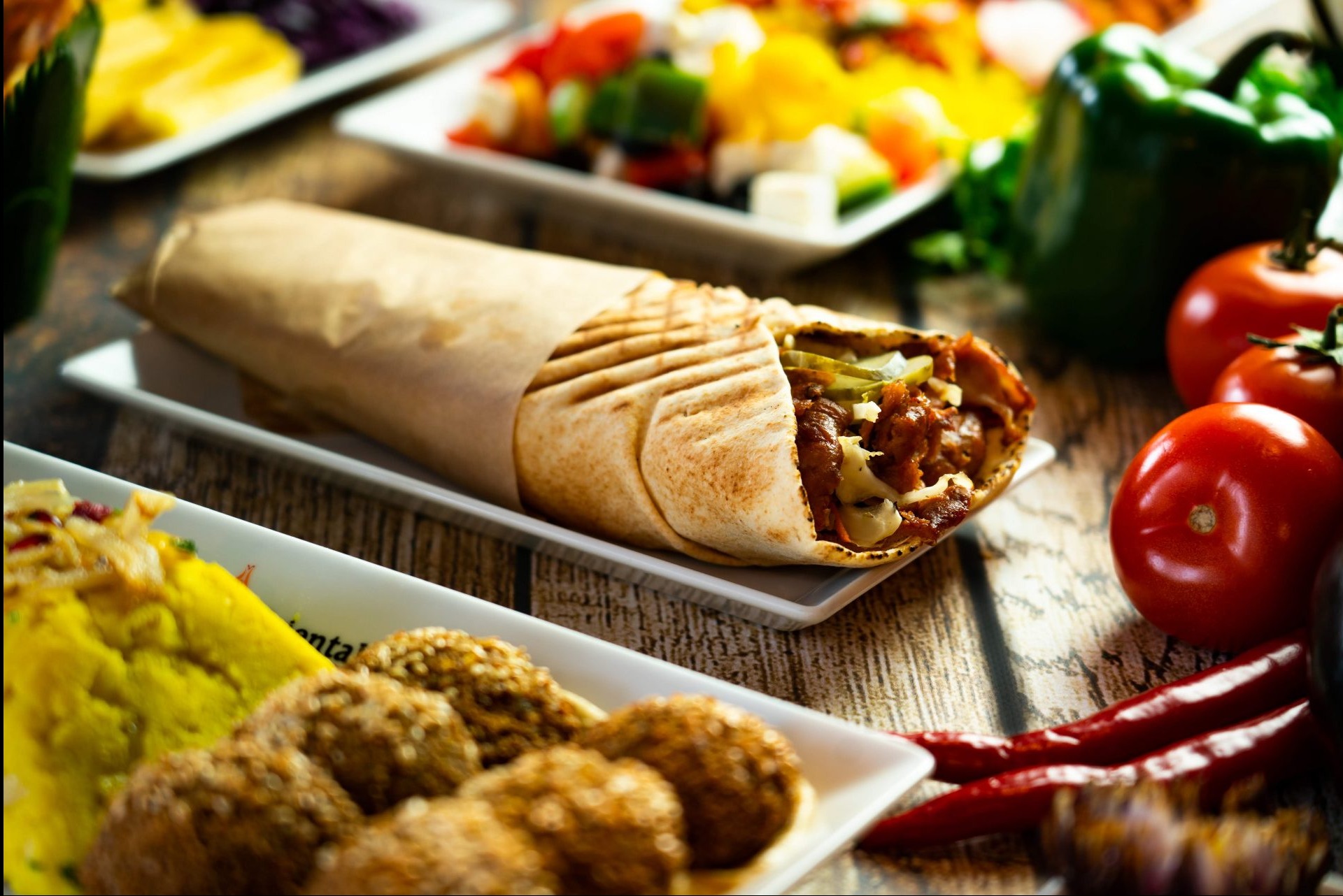 Shawarma w papierze na białym talerzyku, obok pomidory, papryczki chili, falafel i sałatki na drewnianym stole.