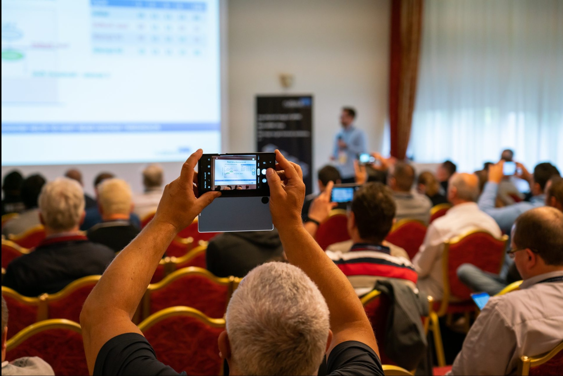 Osoba fotografuje slajd prezentacji z diagramem na ekranie smartfona podczas konferencji, widoczni inni uczestnicy również robiący zdjęcia.