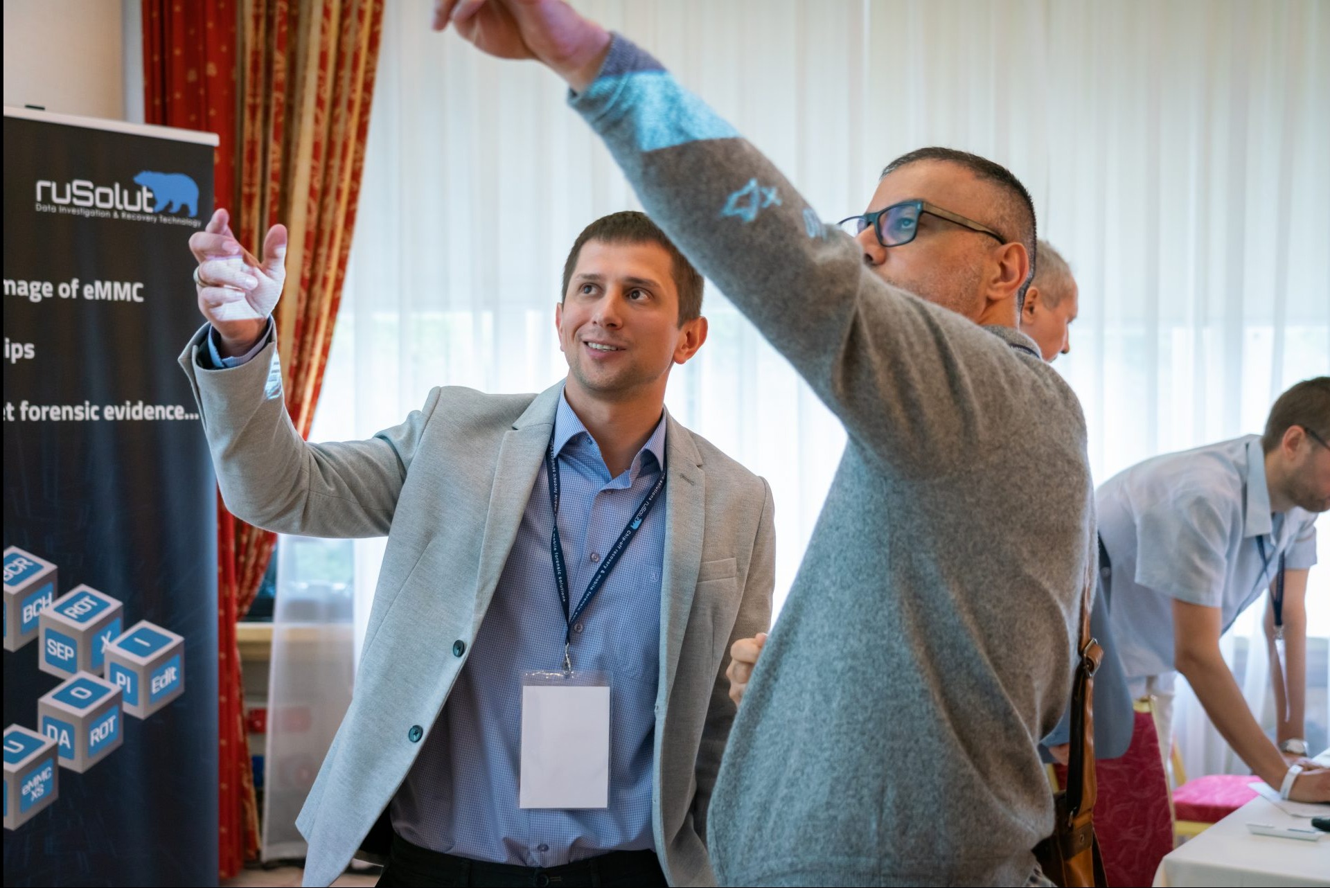 Prezentacja interaktywna: mężczyzna w szarym żakiecie demonstruje cyfrową projekcję na dłoni, obok mężczyzna w okularach obserwuje, w tle baner z logo i kostki z napisami.