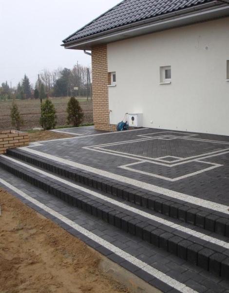 Ułożona kostka brukowa w kolorze grafitowym z białymi akcentami tworząca schody i taras przy budynku z białymi ścianami i ceglanym narożnikiem.