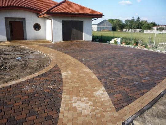 Ułożona kostka brukowa w dwóch odcieniach brązu tworzy ścieżkę i podjazd przed domem z czerwoną dachówką, widok na trawnik w tle.