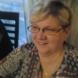 Portret uśmiechniętej kobiety w okularach i wzorzystej bluzce, z widocznym fragmentem krzesła i zasłony w tle.