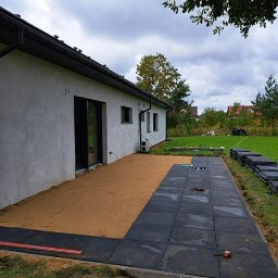 HOMELIKE - Częściowo ułożona, czarna kostka brukowa na piaskowym podłożu przylegająca do ściany budynku. W tle trawnik i drzewa. Widoczne narzędzia i materiały budowlane.