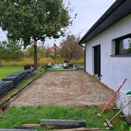 HOMELIKE - Przygotowany teren pod kostkę brukową przy domu. Widoczne obrzeża, podsypka piaskowa, stos kostki i narzędzia. Tło: drzewo, trawa, zabawki.