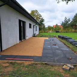 HOMELIKE - Częściowo ułożony taras z ciemnej kostki. Widoczny piasek, poziomica i stos kostki. Dom z czarną stolarką okienną w tle, trawnik i drzewa.
