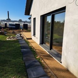 HOMELIKE - Układanie ścieżki z kwadratowych płyt betonowych przy domu z dużym oknem przesuwnym. Ścieżka częściowo ułożona, widoczne przygotowane podłoże z piasku.