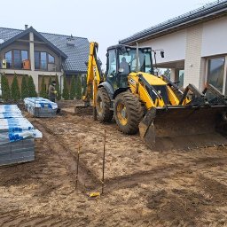 HOMELIKE - Żółta koparko-ładowarka JCB na placu budowy, przygotowuje teren pod kostkę brukową. W tle ułożone palety z kostką i domy.