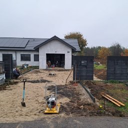 HOMELIKE - Etap prac: przygotowanie podłoża pod kostkę brukową z użyciem zagęszczarki. W tle widoczny fragment ogrodzenia z betonowych bloczków i dom jednorodzinny.