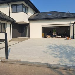 HOMELIKE - Jasnoszara kostka brukowa na podjeździe nowoczesnego domu z garażem. Widoczny fragment ulicy i chodnika. Osoba w tle i narzędzia w otwartym garażu.