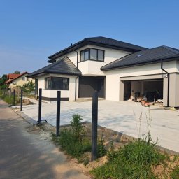 HOMELIKE - Nowoczesny dom jednorodzinny z jasną elewacją i antracytową kostką brukową na podjeździe. W tle widoczne słupki pod ogrodzenie panelowe. Słoneczny dzień.