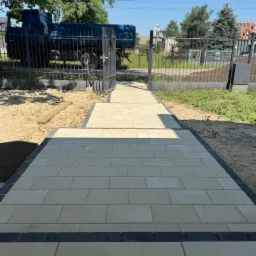 Jasna kostka brukowa z ciemną obwódką prowadzi do metalowej bramy, za którą stoi niebieska ciężarówka. Ujęcie z dołu podkreśla nową nawierzchnię.