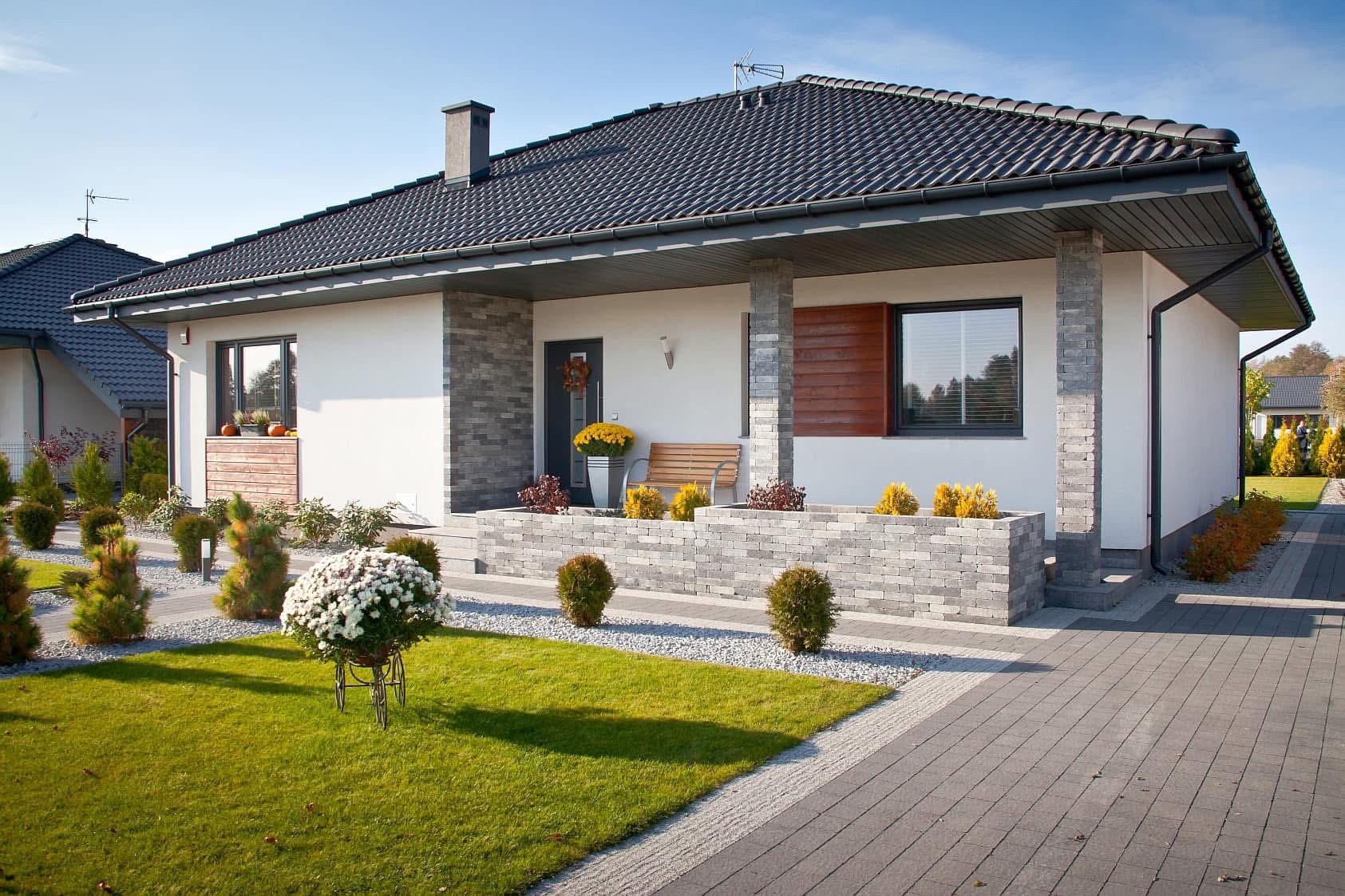 Nowoczesny dom jednorodzinny z antracytową dachówką, jasną elewacją i podjazdem z szarej kostki brukowej. Dekoracyjne rośliny i kwiaty zdobią ogród.