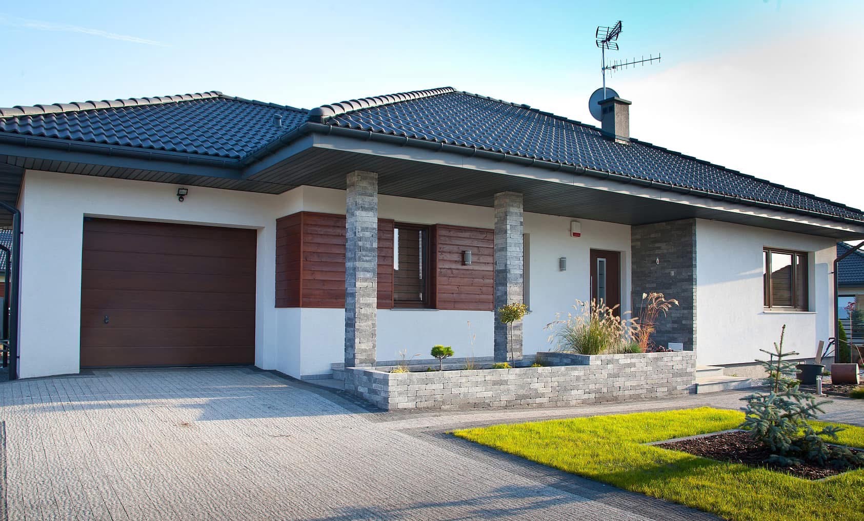 Nowoczesny dom jednorodzinny z szarą kostką brukową na podjeździe i chodniku, widoczny fragment trawnika i elementy architektury ogrodowej.