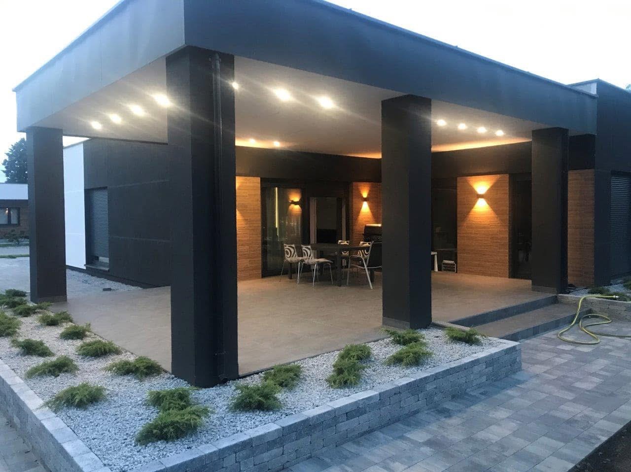 Nowoczesny taras z kostki brukowej w kolorze szarym, otoczony rabatą z kamyczków i niskich krzewów, z widocznym zadaszeniem wspartym na czarnych kolumnach i oświetleniem punktowym.