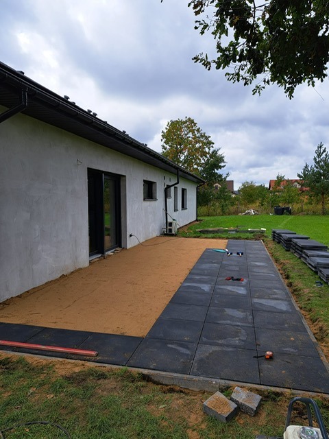 Częściowo ułożona, czarna kostka brukowa na piaskowym podłożu przylegająca do ściany budynku. W tle trawnik i drzewa. Widoczne narzędzia i materiały budowlane.