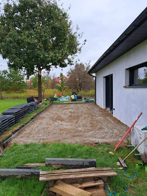 Przygotowany teren pod kostkę brukową przy domu. Widoczne obrzeża, podsypka piaskowa, stos kostki i narzędzia. Tło: drzewo, trawa, zabawki.