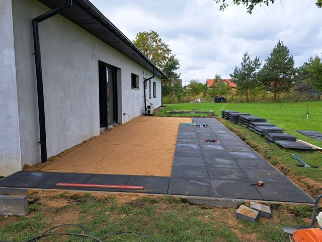 Częściowo ułożony taras z ciemnej kostki. Widoczny piasek, poziomica i stos kostki. Dom z czarną stolarką okienną w tle, trawnik i drzewa.