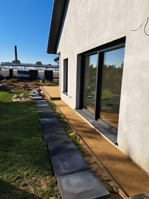 Układanie ścieżki z kwadratowych płyt betonowych przy domu z dużym oknem przesuwnym. Ścieżka częściowo ułożona, widoczne przygotowane podłoże z piasku.