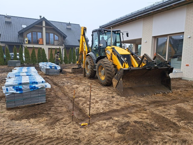 Żółta koparko-ładowarka JCB na placu budowy, przygotowuje teren pod kostkę brukową. W tle ułożone palety z kostką i domy.