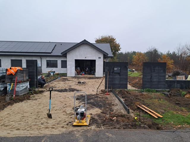 Etap prac: przygotowanie podłoża pod kostkę brukową z użyciem zagęszczarki. W tle widoczny fragment ogrodzenia z betonowych bloczków i dom jednorodzinny.