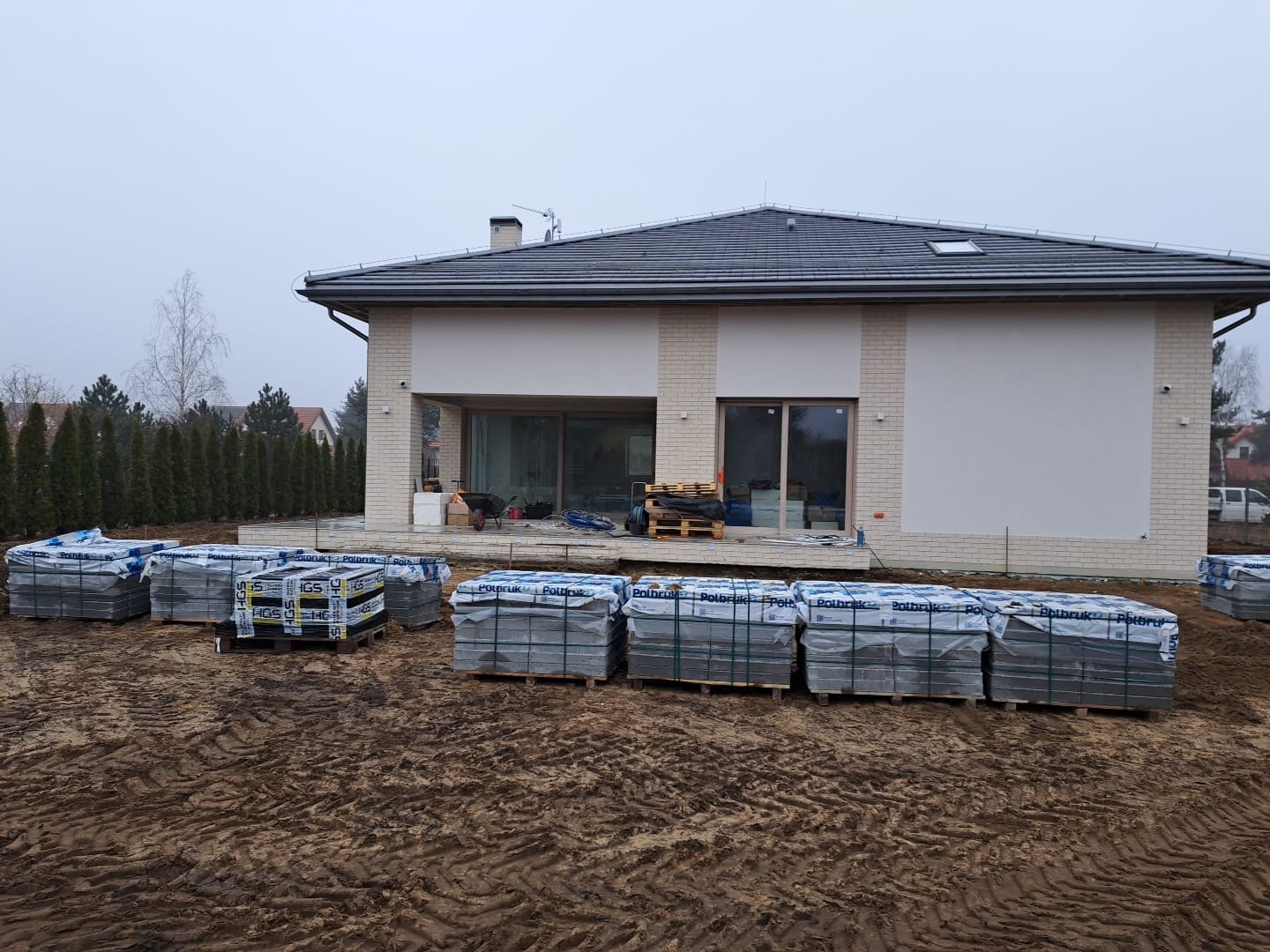 Pakiety kostki brukowej Polbruk na paletach, przygotowane na placu budowy przed nowoczesnym domem z beżowej cegły, w pochmurny dzień.