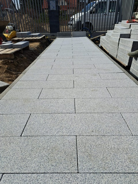 Ułożony chodnik z szarych, prostokątnych płyt, prowadzący do bramy z metalowymi prętami. W tle widoczny samochód dostawczy i przygotowane płyty.