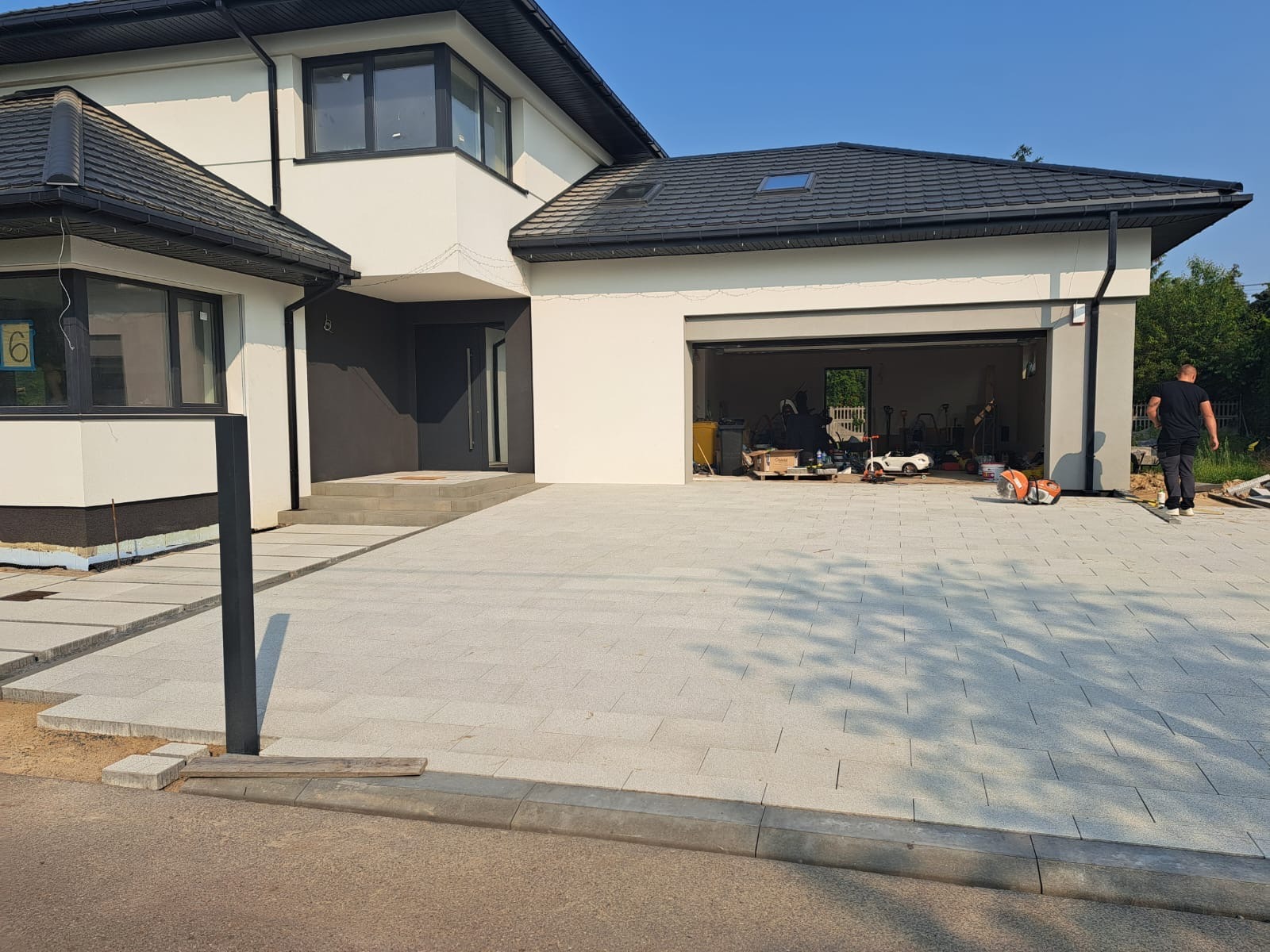 Jasnoszara kostka brukowa na podjeździe nowoczesnego domu z garażem. Widoczny fragment ulicy i chodnika. Osoba w tle i narzędzia w otwartym garażu.