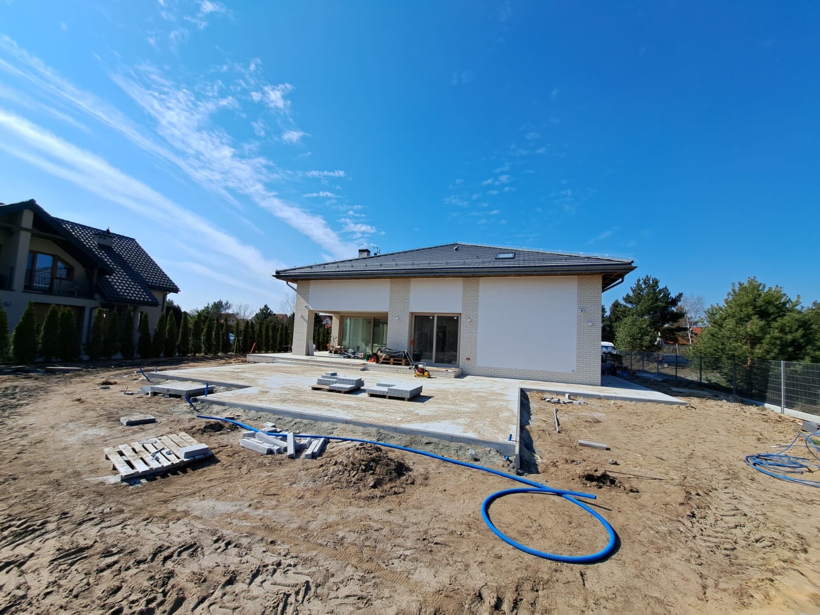 Budowa tarasu z kostki brukowej przy nowoczesnym domu jednorodzinnym w słoneczny dzień. Widoczne materiały budowlane i teren przygotowany pod dalsze prace.
