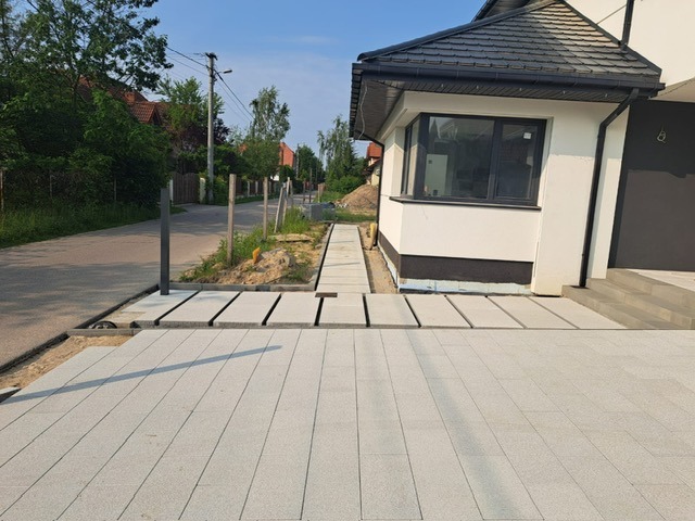 Jasnoszara kostka brukowa ułożona przed nowoczesnym domem z białymi ścianami i ciemną stolarką okienną. Widoczny fragment chodnika i słupka ogrodzeniowego.