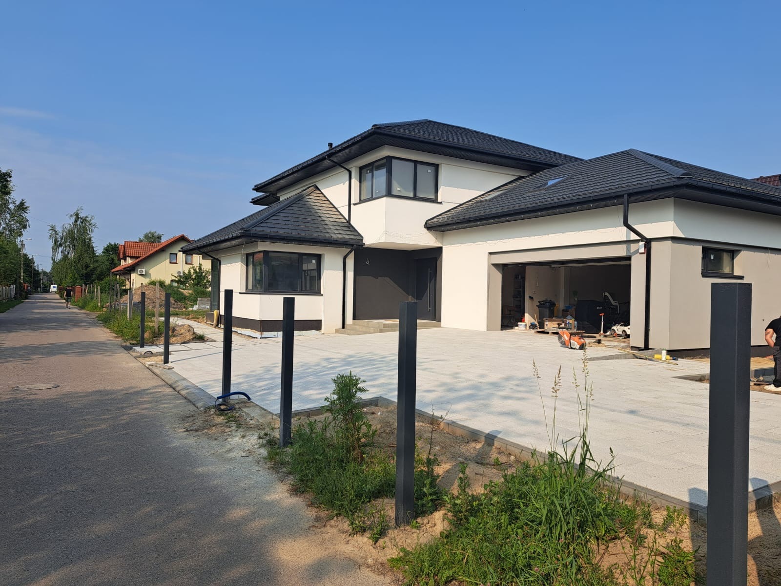 Nowoczesny dom jednorodzinny z jasną elewacją i antracytową kostką brukową na podjeździe. W tle widoczne słupki pod ogrodzenie panelowe. Słoneczny dzień.