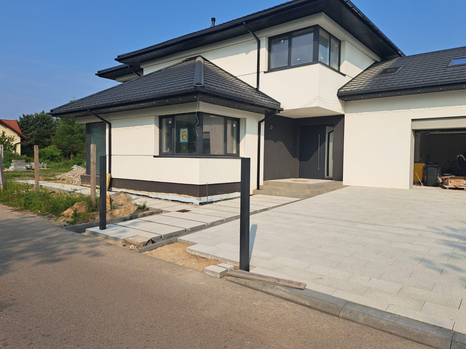 Nowoczesny dom z jasną elewacją i ciemnym dachem. Ułożona jasna kostka brukowa na podjeździe i chodniku. Widoczne słupki pod ogrodzenie. Częściowo wykonane prace brukarskie.