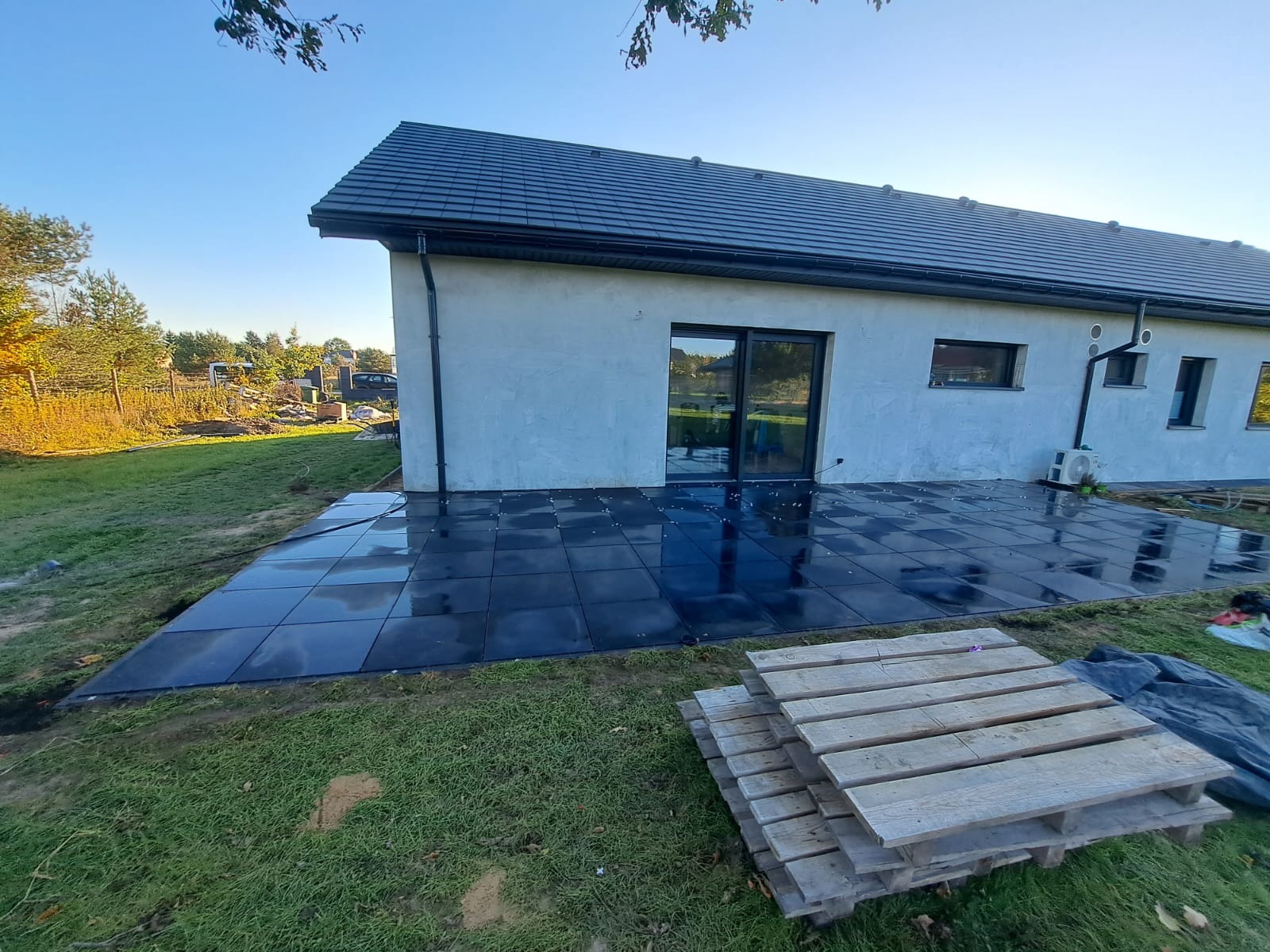 Nowoczesny taras z ciemnej kostki przy domu z jasną elewacją i czarnymi oknami. Widoczna paleta z drewnem. Świeżo ułożona kostka.