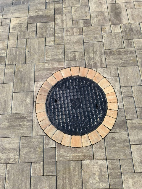 Szara kostka brukowa z okrągłą studzienką, obłożoną jasnymi cegłami, tworzącą estetyczne wykończenie. Widok z góry.
