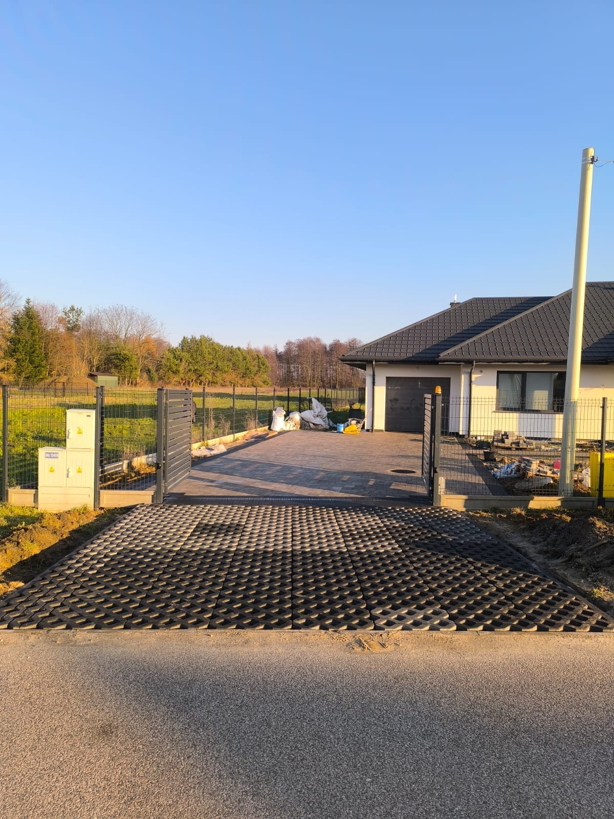 Świeżo ułożona, ażurowa kostka brukowa prowadząca do posesji z szarą bramą i ogrodzeniem panelowym w tle, w słoneczny dzień.