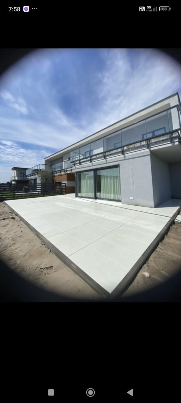 Nowoczesny taras z jasnych płyt na tle domu z dużymi oknami i szklanymi balustradami. Widoczny fragment ogrodzenia panelowego. Całość skąpana w naturalnym świetle słonecznym.