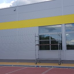 Firma Wielobranżowa EMPE - Szara elewacja budynku przemysłowego z żółtym pasem, widoczne okna z odbijającym się niebem i rusztowanie na kółkach.