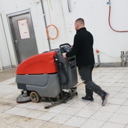 Firma Wielobranżowa EMPE - Mężczyzna obsługuje maszynę do czyszczenia podłóg w pomieszczeniu przemysłowym z białymi ścianami i szarą podłogą w kratkę. Widoczne ślady po czyszczeniu.