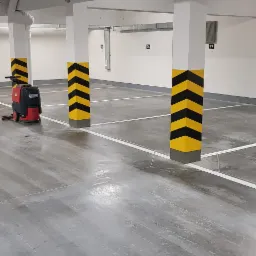 Maszyna do czyszczenia podłóg w garażu podziemnym, widoczne żółto-czarne pasy na kolumnach i białe linie wyznaczające miejsca parkingowe.