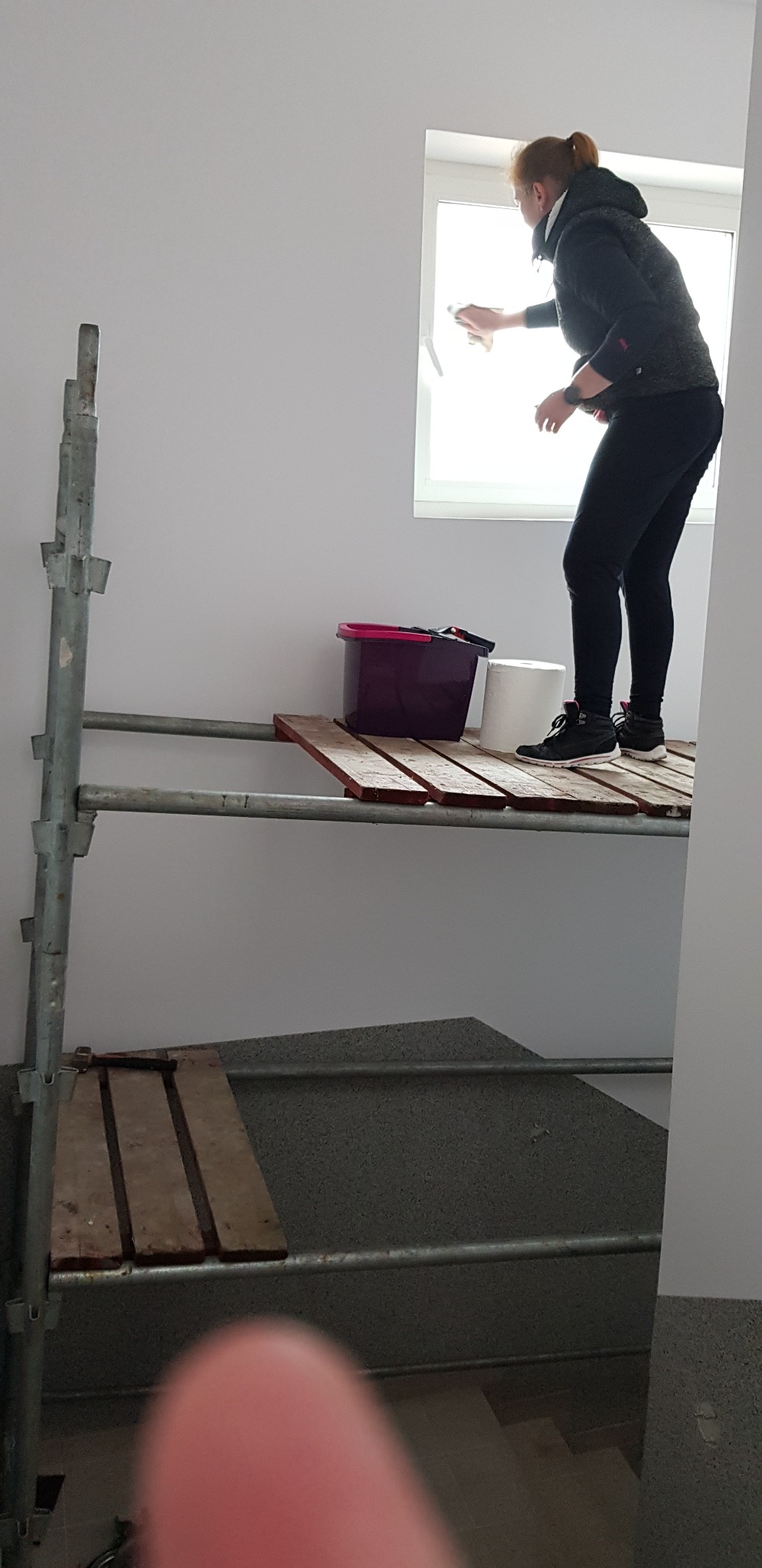 Kobieta w czarnej kurtce i legginsach czyści okno stojąc na prowizorycznym rusztowaniu w jasnym pomieszczeniu, obok wiadro i rolka papieru.