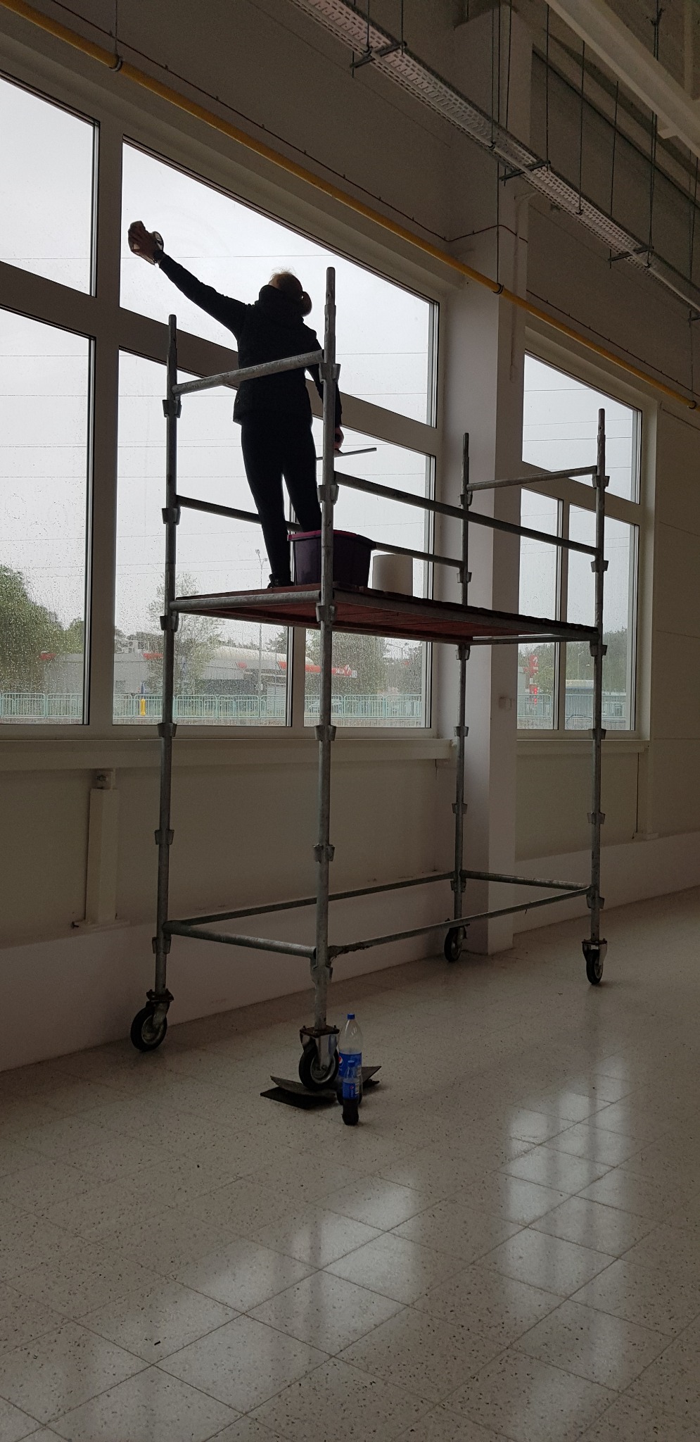 Pracownik na rusztowaniu myje okna w dużym pomieszczeniu; widoczne wiadro, ściereczka i butelka wody.