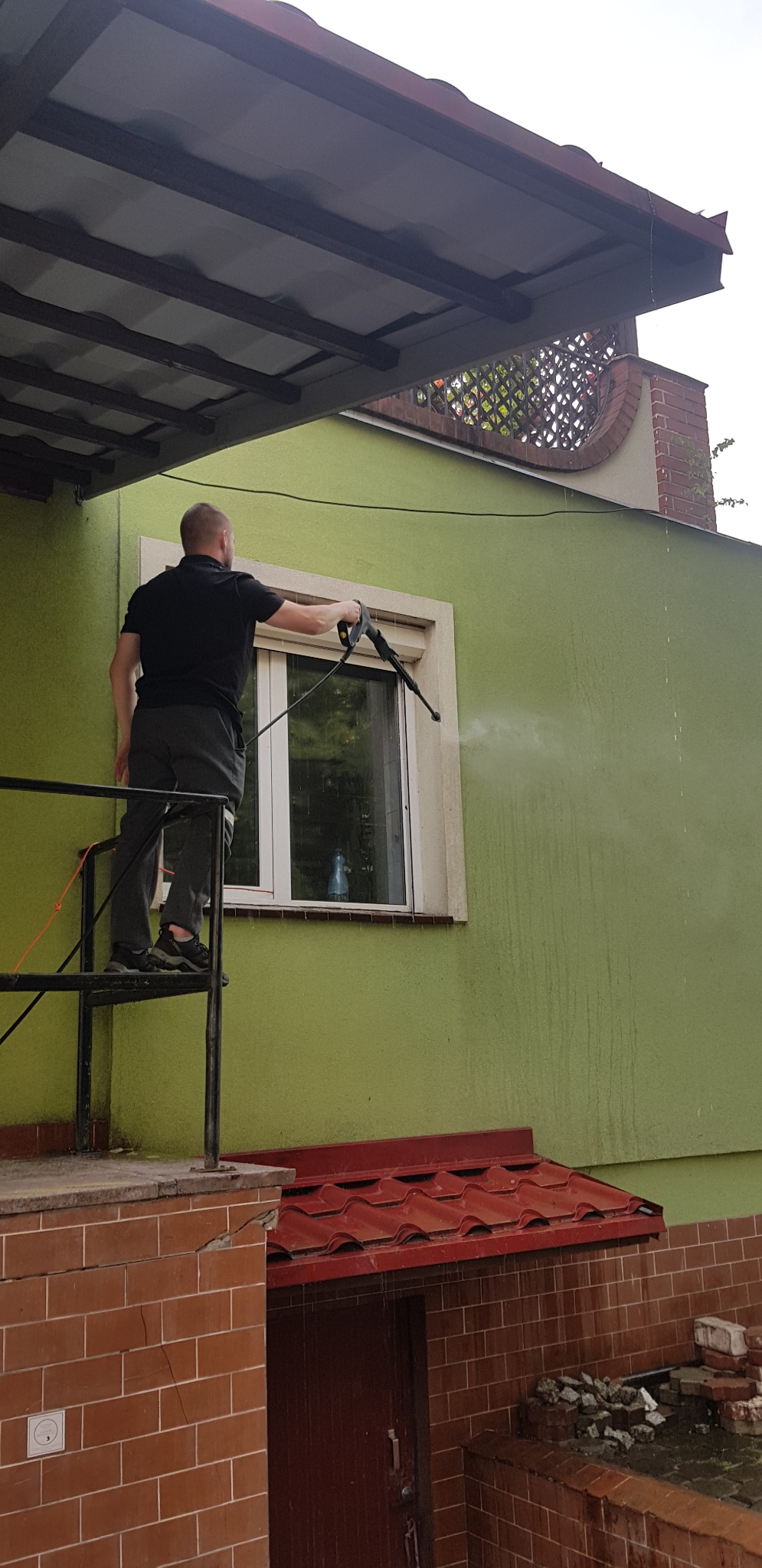 Mężczyzna na rusztowaniu myje elewację domu zieloną myjką ciśnieniową, w tle okno z roletami i fragment dachu.